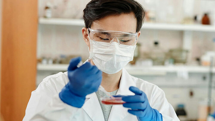 Ein Wissenschaftler, der einen Laborkittel, eine Schutzbrille, eine Maske und blaue Handschuhe trägt, untersucht eine Petrischale mit einer Pipette in einer Laborumgebung, um Krankheiten wie ME/CFS zu erforschen.