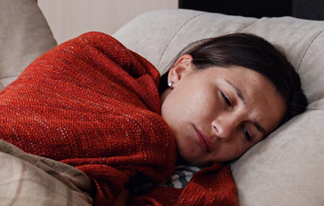 Eine Frau liegt auf einer Couch, den Kopf auf einem Kissen, eingewickelt in eine rote Decke, und sieht müde oder unwohl aus - möglicherweise zeigt sie Symptome einer Krankheit wie ME/CFS.