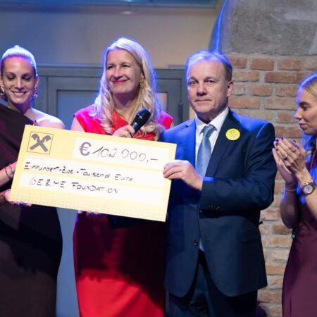 Six formally dressed people stand on a stage, smiling and applauding, while two of them hold an oversized check for €100,000 made out to the SLBENE Foundation, with a brick wall in the background.