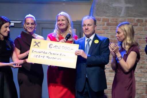Six formally dressed people stand on a stage, smiling and applauding, while two of them hold an oversized check for €100,000 made out to the SLBENE Foundation, with a brick wall in the background.