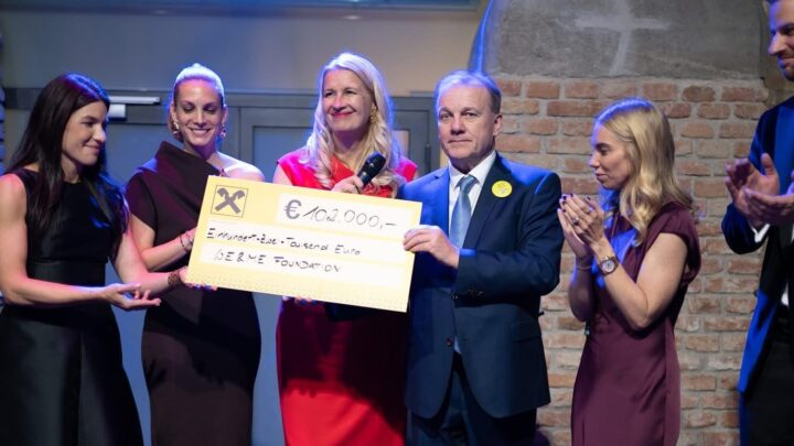 Six formally dressed people stand on a stage, smiling and applauding, while two of them hold an oversized check for €100,000 made out to the SLBENE Foundation, with a brick wall in the background.