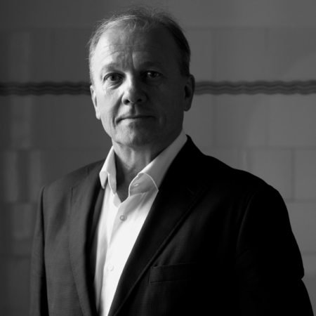 image of Schwarz-Weiß-Porträt eines Mannes mittleren Alters in Anzug und offenem Hemd, der vor einer Kachelwand steht, die von einer Seite dramatisch beleuchtet wird.