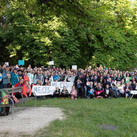 A large group of people pose outdoors in a park, many holding signs that read ME/CFS. Trees and greenery surround them, and a wooden bench and a standing banner are visible in the foreground. 