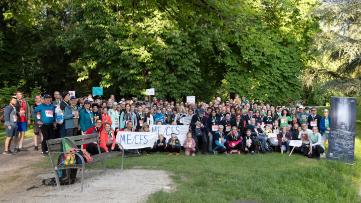 A large group of people pose outdoors in a park, many holding signs that read ME/CFS. Trees and greenery surround them, and a wooden bench and a standing banner are visible in the foreground. 