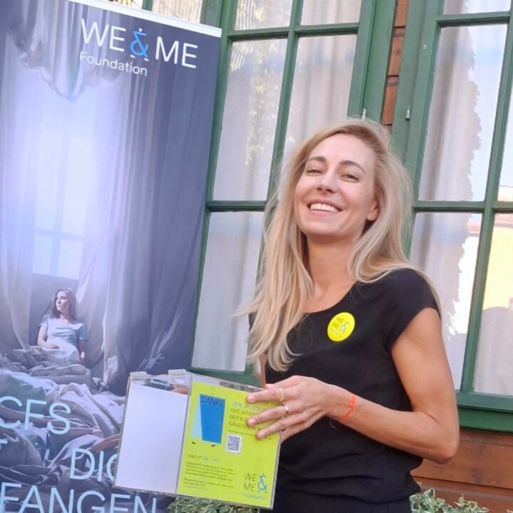 Eine lächelnde Frau mit langen blonden Haaren steht neben einem Banner der WE & ME Foundation und hält Informationsmaterial, das den Gemeinsamen Geist hervorhebt, draußen in der Nähe eines Holzgebäudes mit grün umrahmten Fenstern.