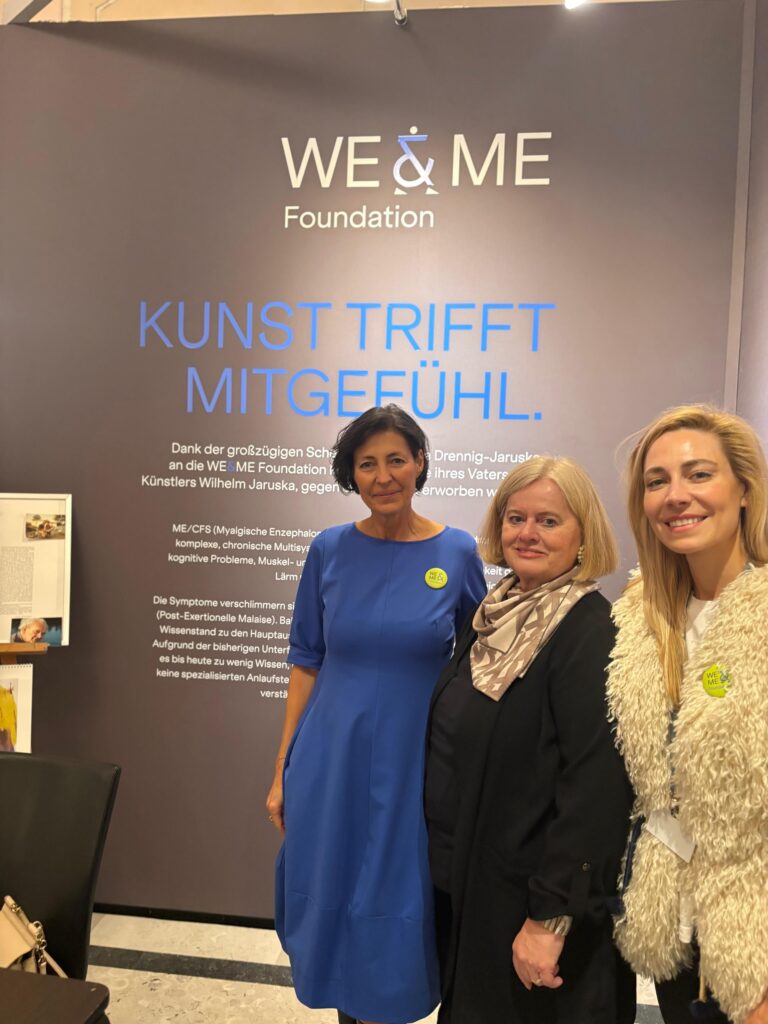 Three women stand smiling in front of a display by the WE & ME Foundation with the German text “Art meets compassion.” Two of the women are wearing light-colored jackets, and one is wearing a blue dress. 