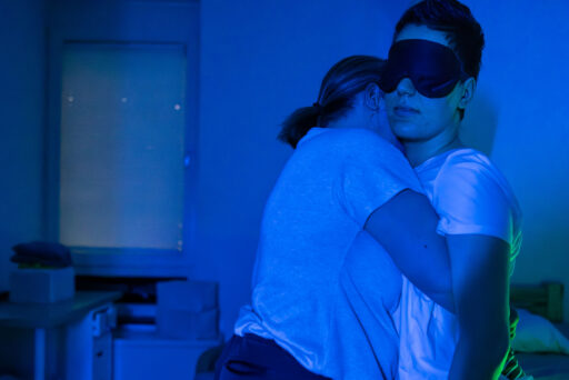 Zwei Personen umarmen sich in einem schwach beleuchteten Raum; eine Person trägt eine Augenmaske und steht neben einem Bett, während sich die andere zur Unterstützung anlehnt. Die blaue Beleuchtung schafft eine ruhige, heitere Atmosphäre.
