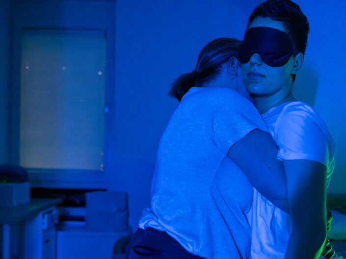 Two people embrace in a dimly lit room; one person wears an eye mask and stands next to a bed, while the other leans against it for support. The blue lighting creates a calm, serene atmosphere. 