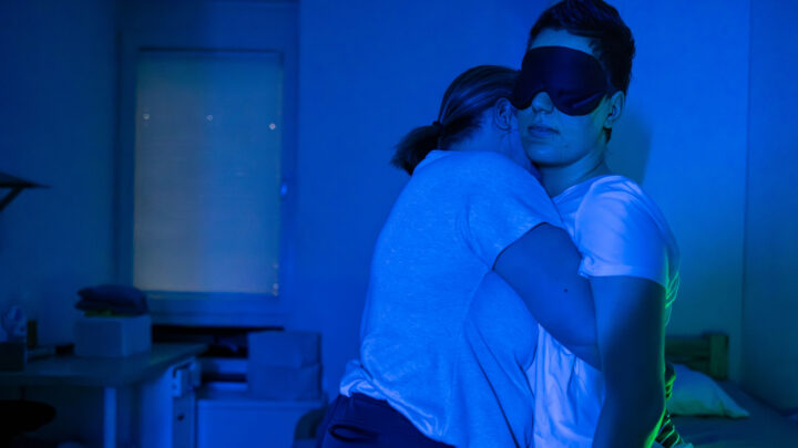 Two people embrace in a dimly lit room; one person wears an eye mask and stands next to a bed, while the other leans against it for support. The blue lighting creates a calm, serene atmosphere. 