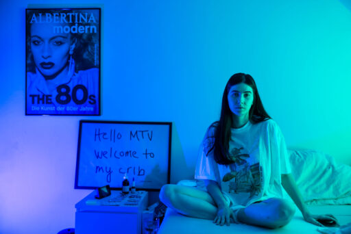 Eine junge Frau sitzt im Schneidersitz auf einem Bett in einem blau-grün beleuchteten Raum. Hinter ihr steht ein Schild mit der Aufschrift "Hello MTV welcome to my crib" und ein Poster einer Frau mit der Aufschrift ALBERTINA modern The 80s. Die Atmosphäre ist entspannt und künstlerisch.