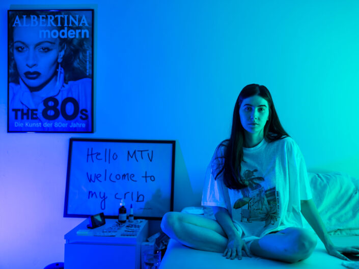 A young woman sits cross-legged on a bed in a room lit in blue-green light. Behind her is a sign that reads “Hello MTV welcome to my crib” and a poster of a woman with the inscription ALBERTINA modern The 80s. The atmosphere is relaxed and artistic.  