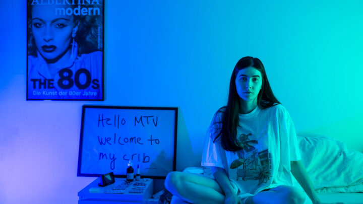 A young woman sits cross-legged on a bed in a room lit in blue-green light. Behind her is a sign that reads “Hello MTV welcome to my crib” and a poster of a woman with the inscription ALBERTINA modern The 80s. The atmosphere is relaxed and artistic.  