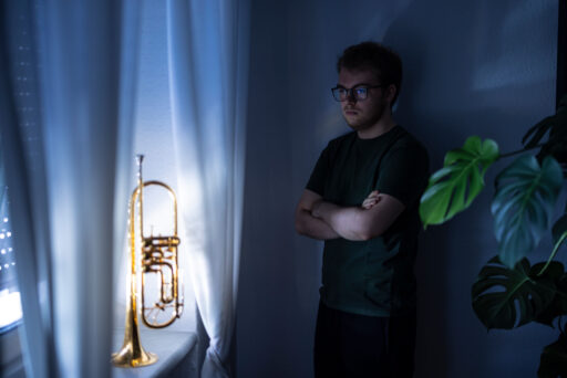 Ein Mann mit Brille steht mit verschränkten Armen in einem schwach beleuchteten Raum und schaut nachdenklich auf eine Messingtrompete auf einem Fensterbrett. In der Nähe befinden sich weiße Vorhänge und eine grüne Blattpflanze.