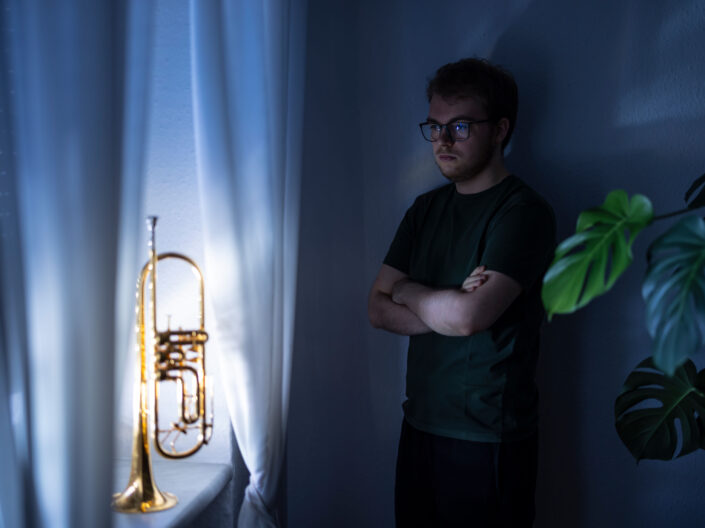 A man wearing glasses stands with his arms crossed in a dimly lit room, looking thoughtfully at a brass trumpet on a windowsill. Nearby are white curtains and a green leafy plant. 