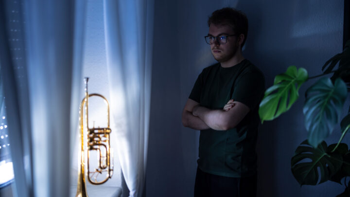 A man wearing glasses stands with his arms crossed in a dimly lit room, looking thoughtfully at a brass trumpet on a windowsill. Nearby are white curtains and a green leafy plant. 