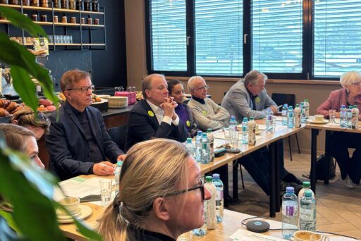 A group of people are sitting around a U-shaped table in a bright room with large windows, holding a meeting. There are water bottles, papers, and coffee cups on the tables. Shelves with glasses and pastries are visible.  