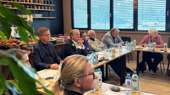 A group of people are sitting around a U-shaped table in a bright room with large windows, holding a meeting. There are water bottles, papers, and coffee cups on the tables. Shelves with glasses and pastries are visible.  