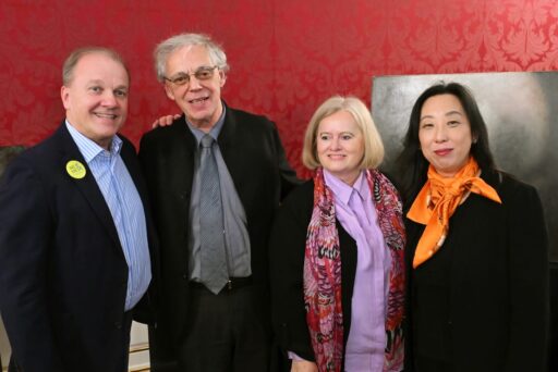 Four adults pose and smile together in a formal room with red patterned wallpaper. On the left are two men in suits, on the right are two women, one wearing a scarf, the other wearing an orange scarf. 