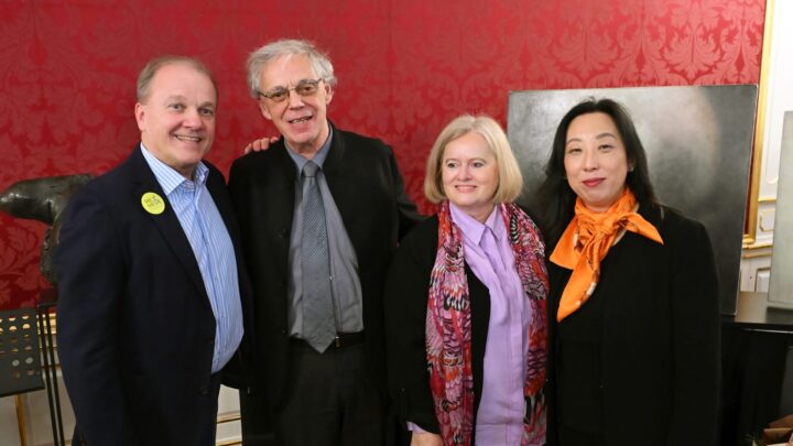 Four adults pose and smile together in a formal room with red patterned wallpaper. On the left are two men in suits, on the right are two women, one wearing a scarf, the other wearing an orange scarf. 
