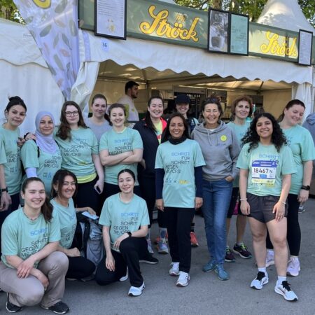 A group of people in matching light blue T-shirts pose and smile in front of a white tent at an outdoor event. Some wear race numbers, others stand or squat together on a sunny day. 