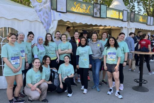 Eine Gruppe von Menschen in passenden hellblauen T-Shirts posiert und lächelt vor einem weißen Zelt bei einer Veranstaltung im Freien. Einige tragen Startnummern, andere stehen oder hocken an einem sonnigen Tag zusammen.