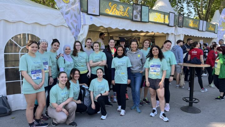 A group of people in matching light blue T-shirts pose and smile in front of a white tent at an outdoor event. Some wear race numbers, others stand or squat together on a sunny day. 