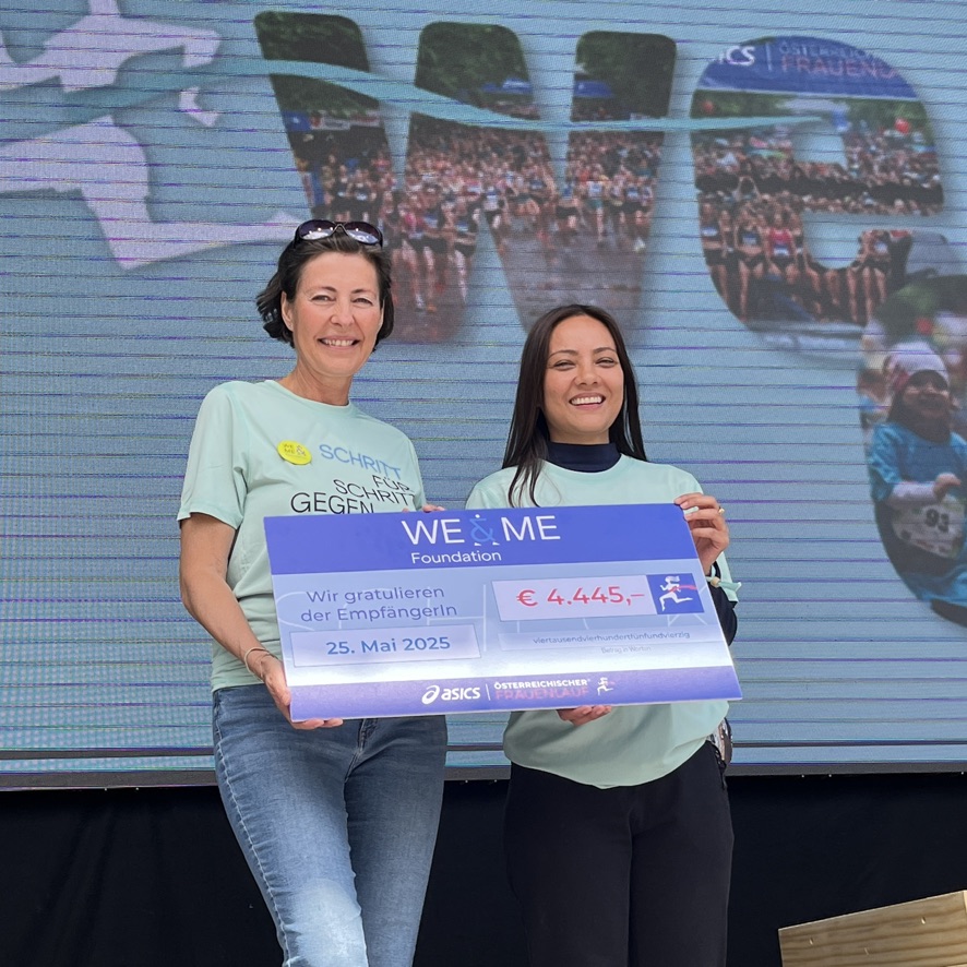 Two women are smiling and holding a large ceremonial cheque for €4,445 from the WE &amp; ME Foundation. They are standing on stage in front of a screen showing graphics of the event. The date on the check is May 25, 2025.  
