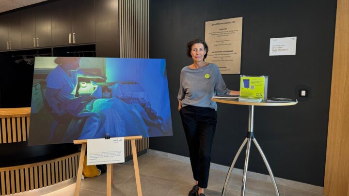 Eine Frau steht neben einem hohen Tisch mit einer kleinen grünen Schachtel und einem Schild. Neben ihr steht eine große Fotografie auf einer Staffelei, die eine medizinische Szene zeigt. Die Umgebung scheint ein moderner Innenraum mit dunklen Wänden und Schränken zu sein.