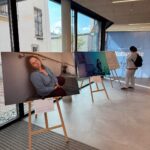 Fotoausstellung in einem modernen Innenraum mit großen Fenstern. Auf Staffeleien sind Porträts ausgestellt; eines zeigt eine Frau mit geschlossenen Augen. Eine Person steht neben einem anderen Porträt und schaut es an. Im Hintergrund ist ein blaues Transparent zu sehen.