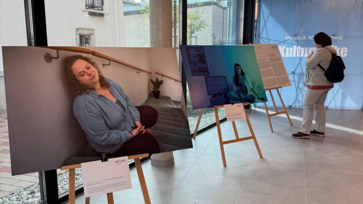 Fotoausstellung in einem modernen Innenraum mit großen Fenstern. Auf Staffeleien sind Porträts ausgestellt; eines zeigt eine Frau mit geschlossenen Augen. Eine Person steht neben einem anderen Porträt und schaut es an. Im Hintergrund ist ein blaues Transparent zu sehen.