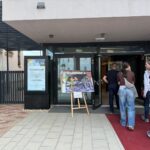 Menschen betreten ein modernes Gebäude mit Glastüren. Am Eingang steht eine Staffelei mit einer großen Karte oder einem Poster auf einem roten Teppich. Links von den Türen ist ein Schild mit Informationen zu sehen.