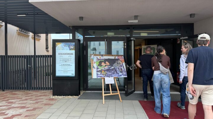 Menschen betreten ein modernes Gebäude mit Glastüren. Am Eingang steht eine Staffelei mit einer großen Karte oder einem Poster auf einem roten Teppich. Links von den Türen ist ein Schild mit Informationen zu sehen.