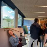 Eine Gruppe von Menschen steht in einem Raum neben einem großen Foto einer Frau, die mit geschlossenen Augen auf einer Treppe sitzt. Der Raum hat große Fenster, moderne Beleuchtung und eine Staffelei mit einem anderen Kunstwerk im Hintergrund.