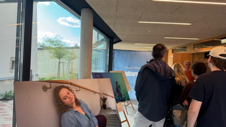 Eine Gruppe von Menschen steht in einem Raum neben einem großen Foto einer Frau, die mit geschlossenen Augen auf einer Treppe sitzt. Der Raum hat große Fenster, moderne Beleuchtung und eine Staffelei mit einem anderen Kunstwerk im Hintergrund.