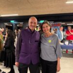 Ein Mann in einer violetten Weste und eine Frau in einem grauen Oberteil lächeln sich bei einer Veranstaltung in einer Halle an, während im Hintergrund Menschen und Plakate zu sehen sind.