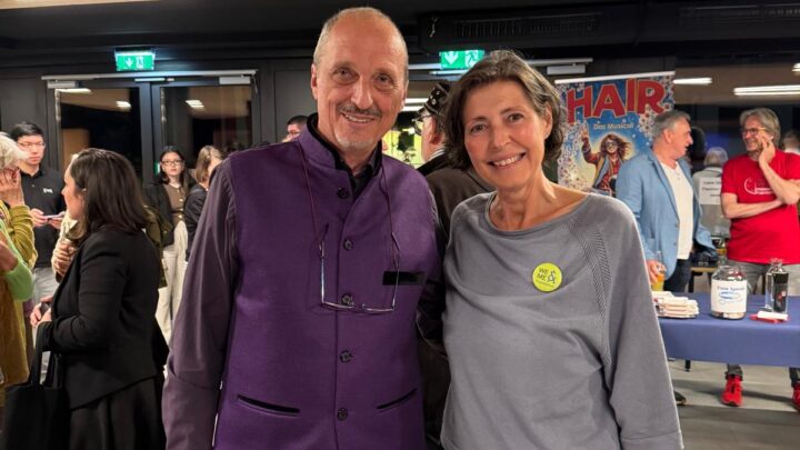 Ein Mann in einer violetten Weste und eine Frau in einem grauen Oberteil lächeln sich bei einer Veranstaltung in einer Halle an, während im Hintergrund Menschen und Plakate zu sehen sind.
