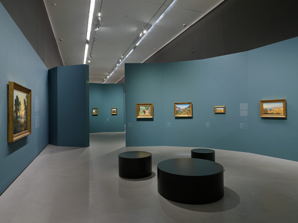 A modern art gallery with curved blue walls displays framed landscape paintings. Round black benches are arranged on the smooth white floor under bright ceiling lights. 