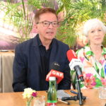 Rudolf Anschober in einem marineblauen Blazer spricht auf einer Pressekonferenz über ME/CFS in Österreich: Vier ehemalige Minister unterstützen die Petition zum PAIS-Aktionsplan. Er sitzt zwischen zwei Frauen, auf dem Tisch stehen Mikrofone, eine Flasche und Unterlagen. Im Hintergrund stehen grüne Pflanzen.