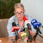 Andrea Kdolsky mit kurzen grauen Haaren und Brille spricht auf einer Pressekonferenz über ME/CFS in Österreich: Vier ehemalige Minister unterstützen die Petition zum PAIS-Aktionsplan. Auf dem Tisch stehen mehrere Mikrofone, Flaschen und Blumen, im Hintergrund sind Pflanzen zu sehen.