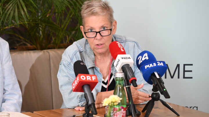 Andrea Kdolsky mit kurzen grauen Haaren und Brille spricht auf einer Pressekonferenz über ME/CFS in Österreich: Vier ehemalige Minister unterstützen die Petition zum PAIS-Aktionsplan. Auf dem Tisch stehen mehrere Mikrofone, Flaschen und Blumen, im Hintergrund sind Pflanzen zu sehen.
