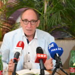 Johannes Rauch mit Brille und hellblauem Blazer spricht auf einer Pressekonferenz über ME/CFS in Österreich: Vier ehemalige Minister unterstützen die Petition zum PAIS-Aktionsplan, wobei er zwischen Mikrofonen, Wasserflaschen und Pflanzen mit den Händen gestikuliert.