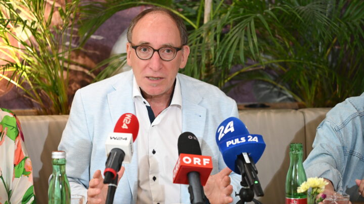 Johannes Rauch mit Brille und hellblauem Blazer spricht auf einer Pressekonferenz über ME/CFS in Österreich: Vier ehemalige Minister unterstützen die Petition zum PAIS-Aktionsplan, wobei er zwischen Mikrofonen, Wasserflaschen und Pflanzen mit den Händen gestikuliert.