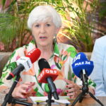 Maria Rauch-Kallat mit kurzen weißen Haaren, die eine bunte Bluse mit Blumenmuster trägt, spricht auf einer Pressekonferenz über ME/CFS in Österreich: Vier ehemalige Minister unterstützen die Petition zum PAIS-Aktionsplan. Im Hintergrund sind grüne Pflanzen zu sehen.