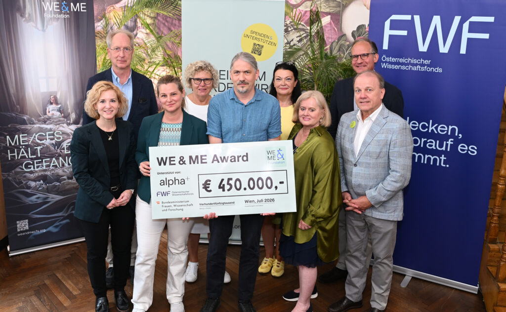 Nine people are standing in a room, holding a large check for €450,000 for the WE&ME Award ME/CFS, which features the logos of the sponsors alpha+, FWF, and WE & ME; event banners can be seen in the background.