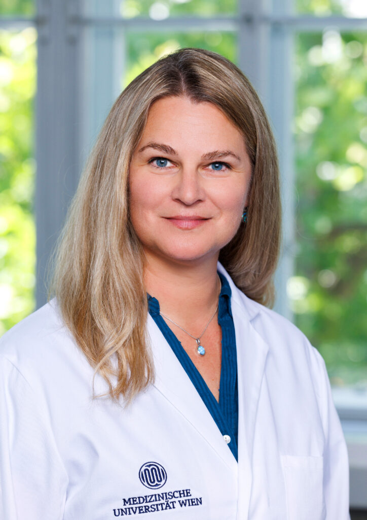 A woman with long blonde hair, wearing a white lab coat and a blue shirt, is standing in front of a window with potted plants behind it. The lab coat bears the logo and name of the Medical University of Vienna, indicating her work in ME/CFS research. 