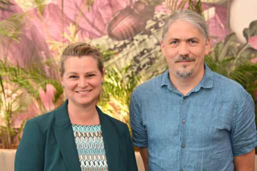 Two adults are standing indoors in front of some plants and a colorful mural; one is wearing a green blazer and a patterned top, and the other is wearing a blue, short-sleeved button-down shirt. Both are smiling and looking at the camera, creating a welcoming atmosphere that reflects their shared passion for ME/CFS research. 