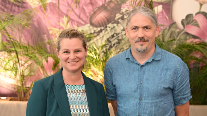 Two adults are standing indoors in front of some plants and a colorful mural; one is wearing a green blazer and a patterned top, and the other is wearing a blue, short-sleeved button-down shirt. Both are smiling and looking at the camera, creating a welcoming atmosphere that reflects their shared passion for ME/CFS research. 