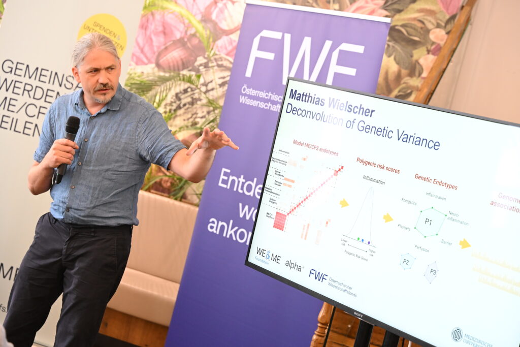 A man is speaking into a microphone and pointing at a large screen showing a scientific presentation on genetic variability and ME/CFS research; banners and a plant can be seen in the background.
