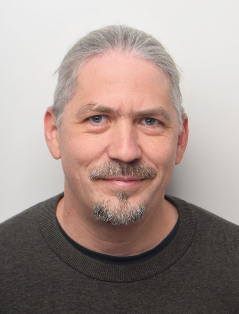 A middle-aged man with fair skin, blue eyes, gray hair combed back, and a short beard and mustache, wearing a dark sweater, smiles at the camera against a plain white background—an expression of determination often seen in people dedicated to ME/CFS research.