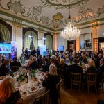 During the charity gala in Kittsee, a large group of people in formal attire sits at round tables in a lavishly decorated hall, listening to the speakers on stage. A magnificent chandelier hangs from an elaborately decorated ceiling. 